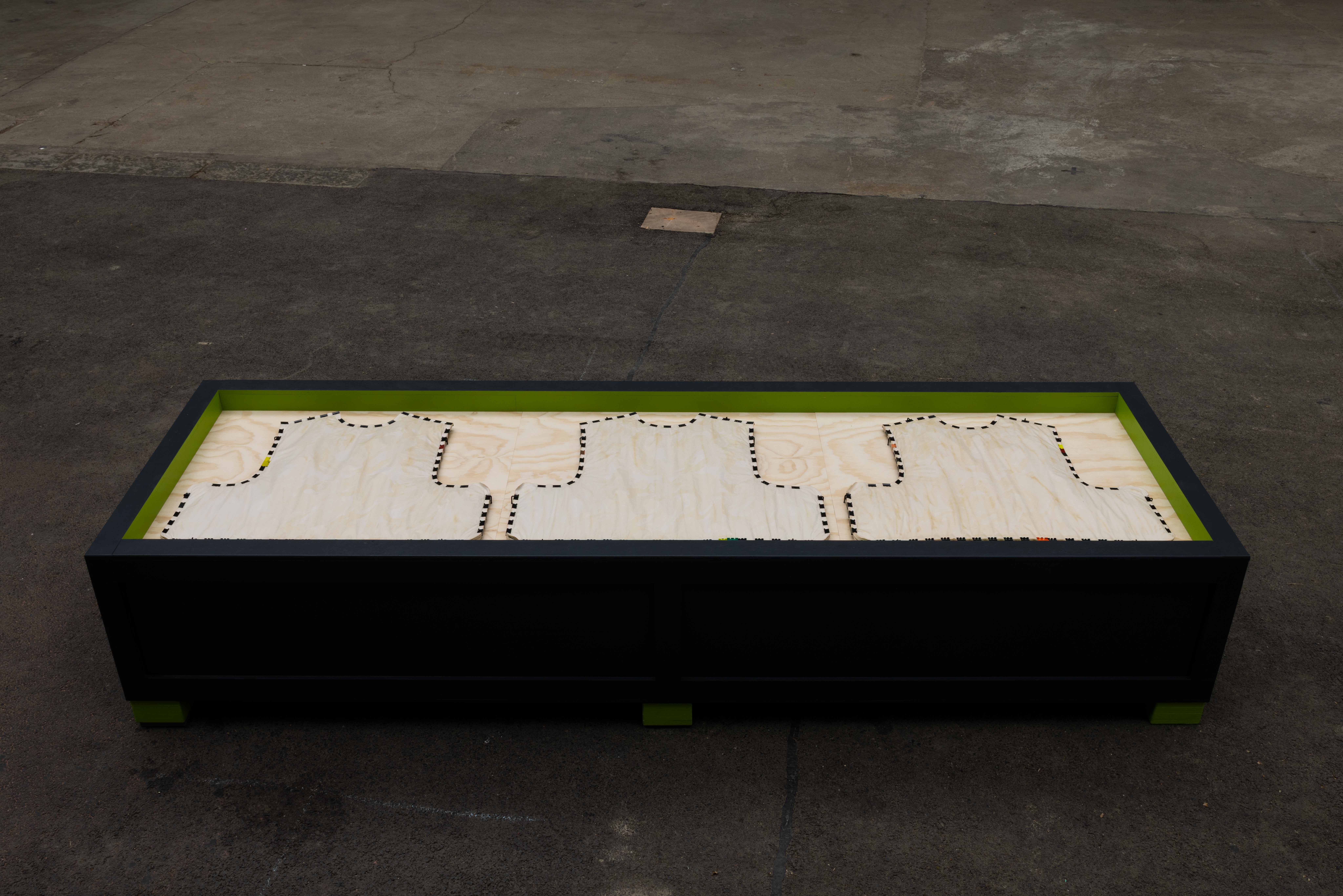Long low black and green wooden crate sculpture on a paved courtyard, with vest-shaped outlines stitched in black onto the plywood base viewed from above.