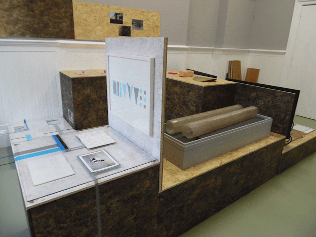 An installation made of different materials has a rectangular shape with different earth-toned colours and white.