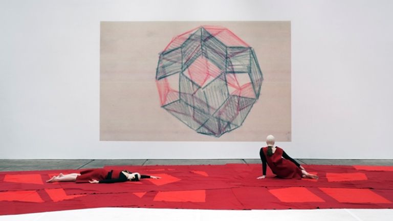 two people dressed in red lying on the floor on top of red pieces of fabric. behind them is a large drawing of a geometric polyhedron/sphere 