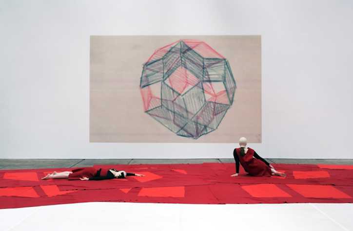 two people dressed in red lying on the floor on top of red pieces of fabric. behind them is a large drawing of a geometric polyhedron/sphere