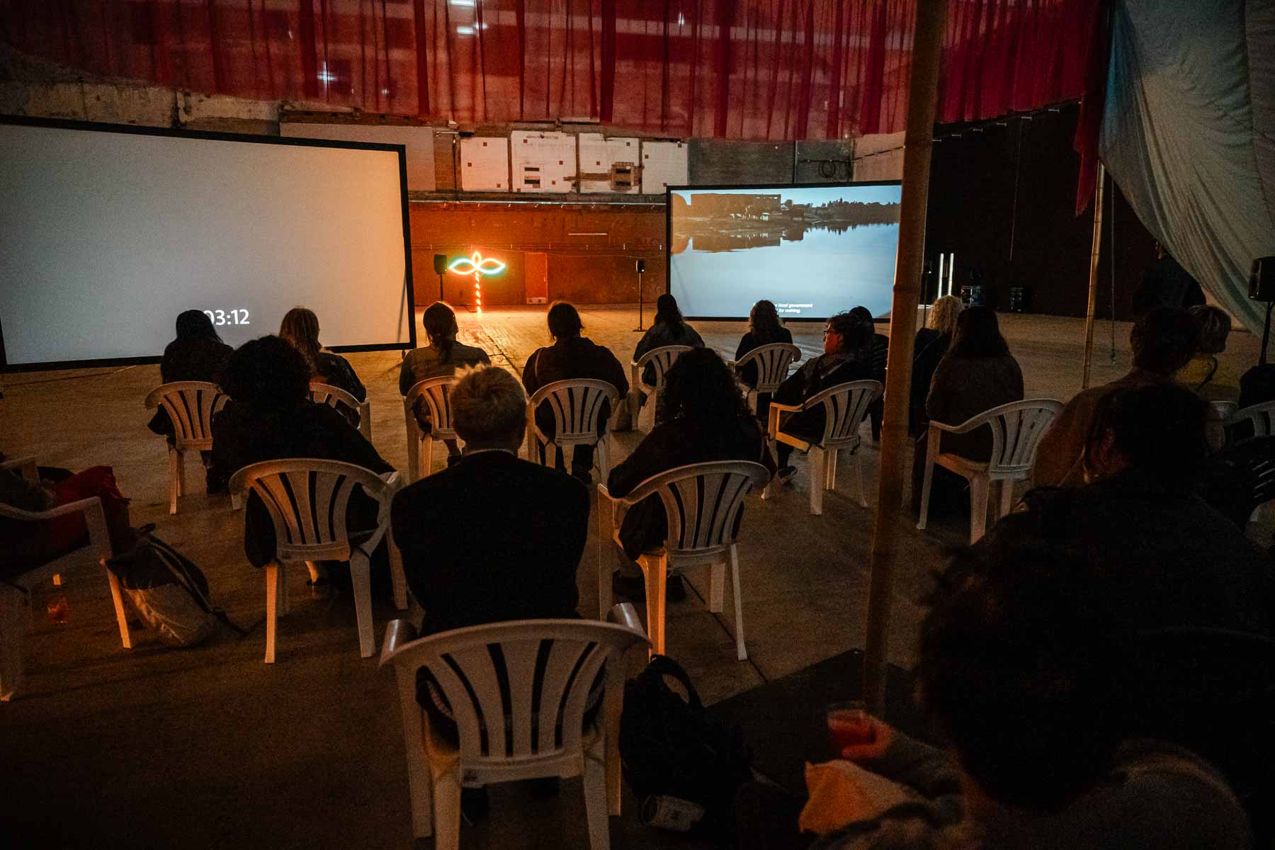 An audience seated on white plastic chairs watches two film projections in a large gallery, with a glowing neon sculpture between the screens.