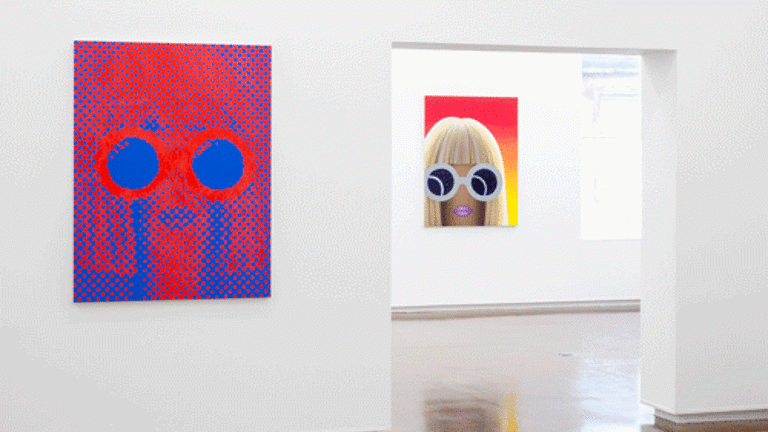 A white walled gallery with two wall-based artworks visible. These are abstracted faces with large round glasses