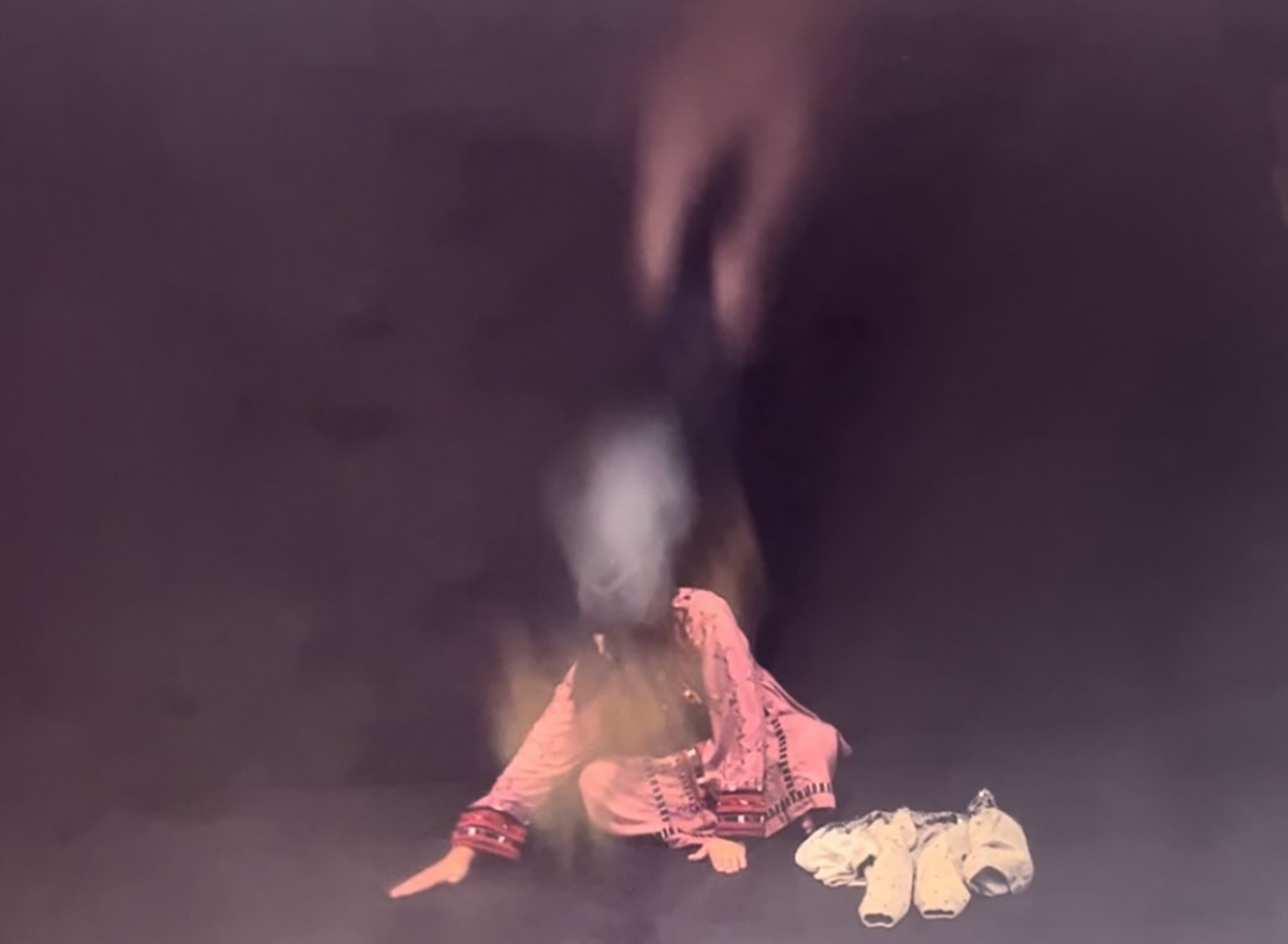 a woman wearing a pink fown with decorative edges sits cross-legges on a stage performing a wiping movement,