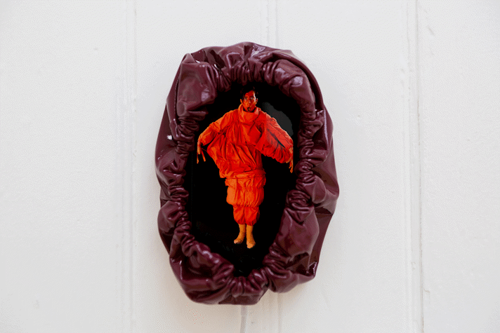 a figure with a painted red face in a red costume stands in blackness with their arms out and mouth in an 'o' shape. Their image is on an iphone mounted on a wall. The iphone is encased in a drawstring burgundy PVC cover