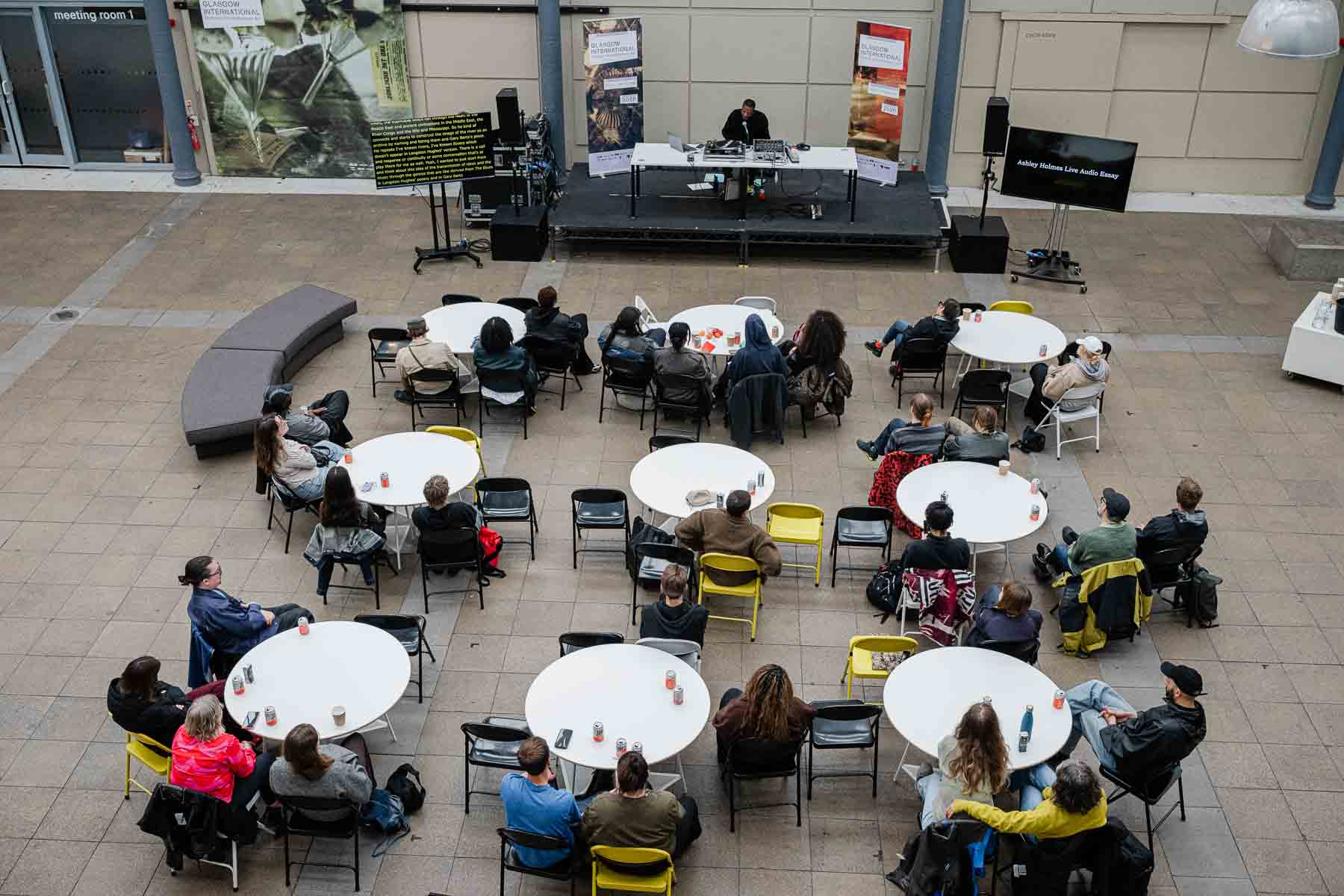 Aerial view of an outdoor courtyard event, with an audience seated at round tables arranged in a loose circle around a small stage. Ashley Holmes stands at the stage performing at a DJ setup, with a screen beside the stage reading "Ashley Holmes Live Audio Essay" and Glasgow International banners in the background.