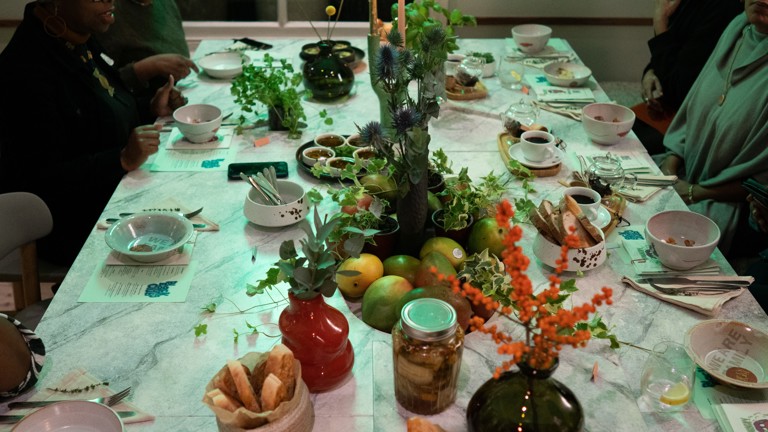 A long table set for a communal meal, decorated with plants, fruit, bread, and ceramic bowls, with people seated around it.