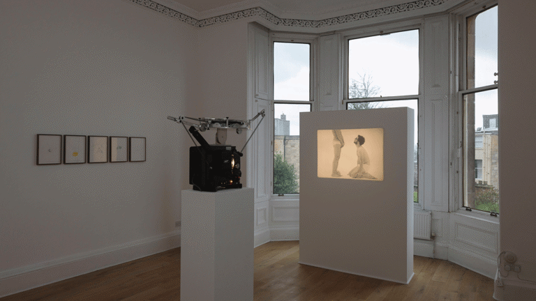 Exhibition view of gallery with framed photographs on the wall and a 16mm film projected onto a free-standing wall. The film images shows 2 figures