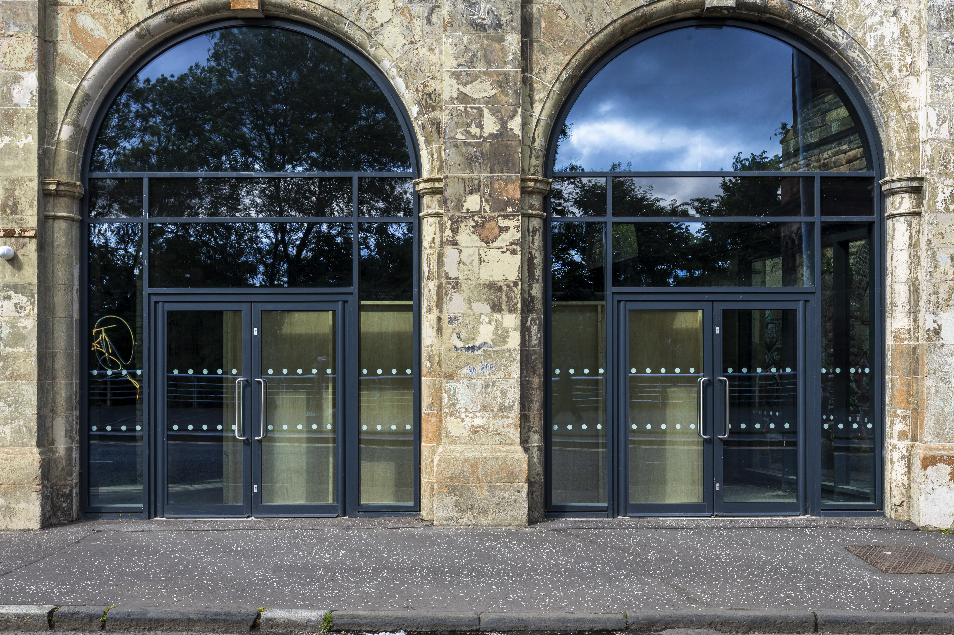 Two arched stone entrances at the Briggait with dark-framed glass doors, reflecting trees and a blue sky, one door marked with yellow graffiti.