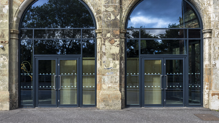 Two arched stone entrances at the Briggait with dark-framed glass doors, reflecting trees and a blue sky, one door marked with yellow graffiti.
