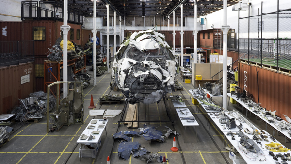 Interior of Tramway 2 with Wreckage of an aircraft reassembled as part of an art installation in an industrial space, with metal fragments, tables displaying parts, and shipping containers in the background.