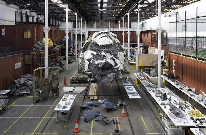 Interior of Tramway 2 with Wreckage of an aircraft reassembled as part of an art installation in an industrial space, with metal fragments, tables displaying parts, and shipping containers in the background.