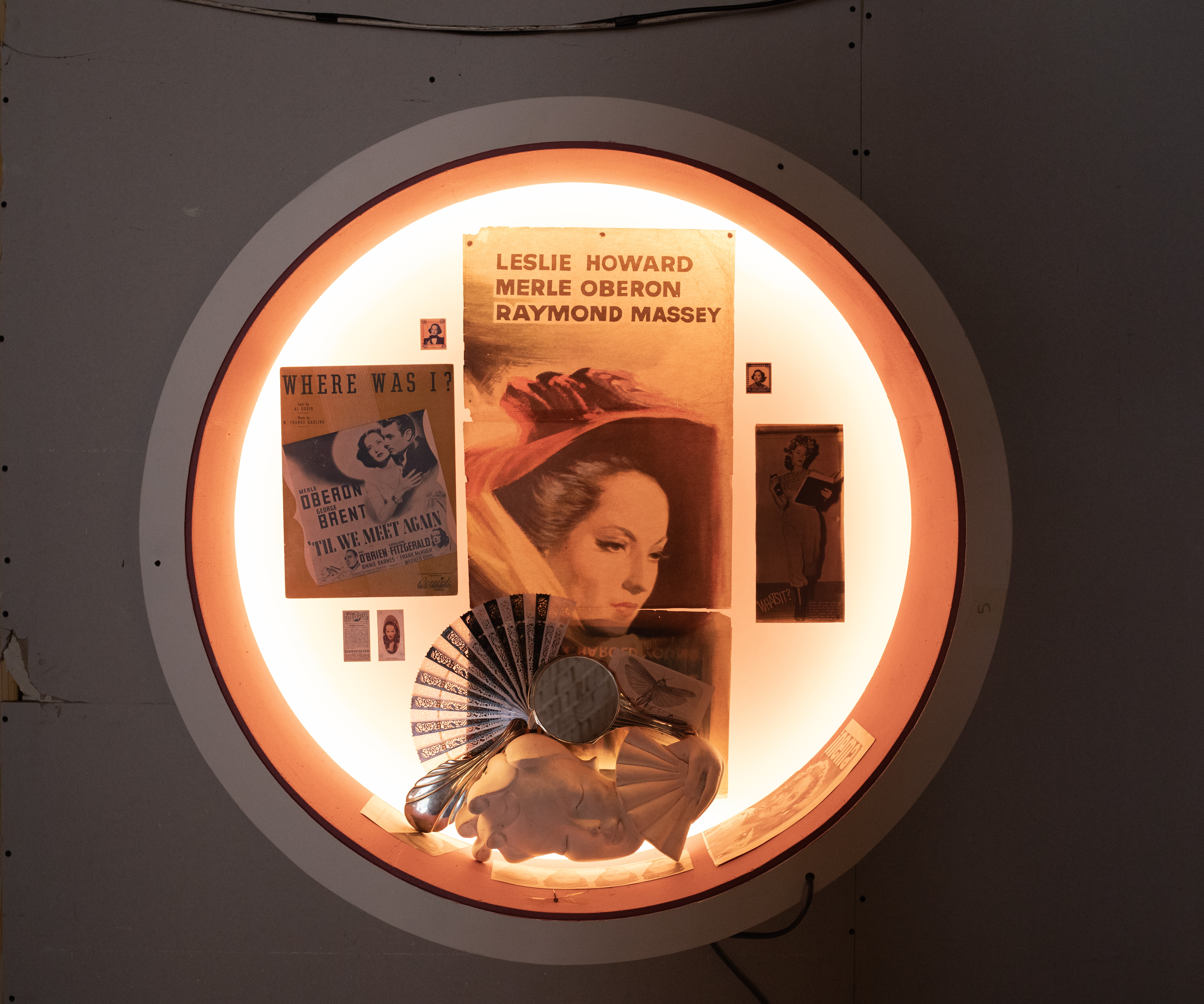 Illuminated circular display featuring vintage movie posters, photos, a fan, mirror, and decorative masks.