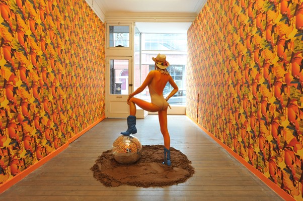 Rear view of a figure wearing a swimming costume with one foot resting on a sphere. In the back ground is a shop window looking on to a street. The walls of the interior have a patterned wallpaper. 