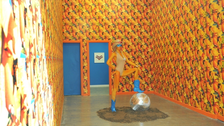 mannequin in a swimsuit and a straw hat standing with one foot on a disco ball in a pile of dirt. the room is covered in bright patterned yellow wallpaper