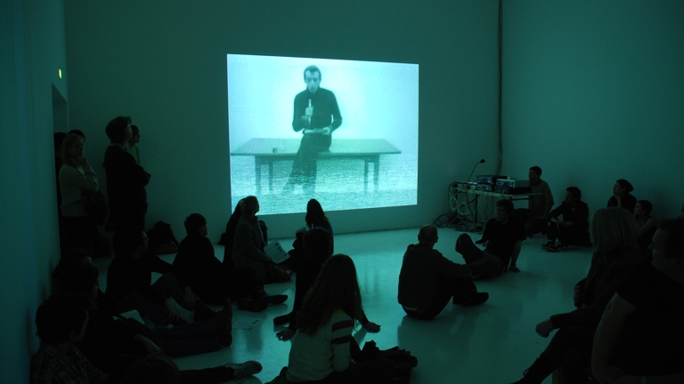 people sat on the floor watching a projection of a person sat on a table