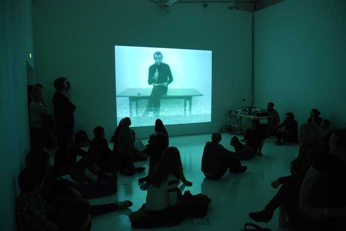people sat on the floor watching a projection of a person sat on a table