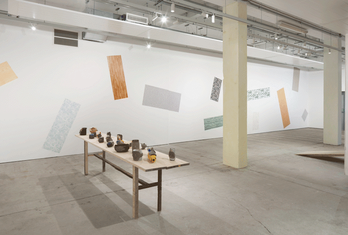 A Wooden table is placed in a long empty hall. There are multiple small clay sculptures placed on the table. The wall on the side of the table has colourful rectangular shapes printed onto it.