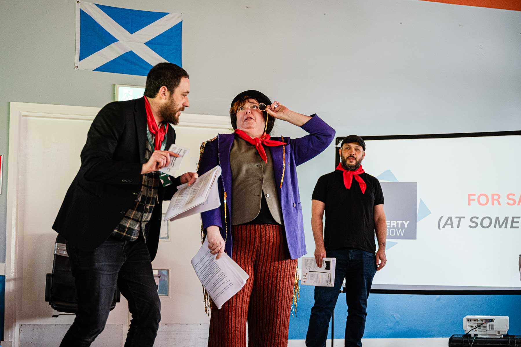 A performer in a purple jacket and monocle gestures upward theatrically, flanked by two other cast members, in front of a screen reading "For sale (at some..." beneath a Saltire flag.
