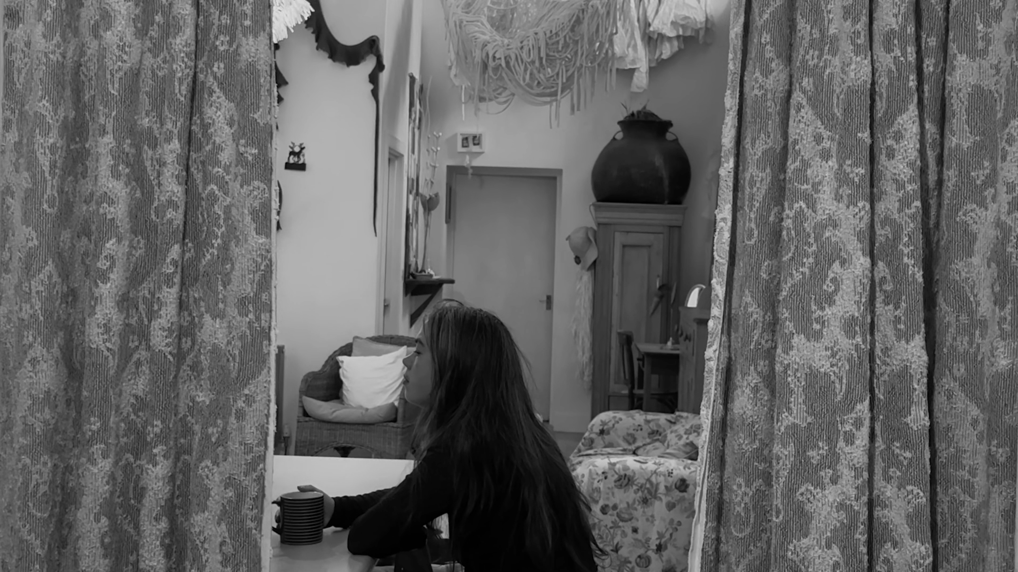 A film still of a person with long hair sitting at a table holding a mug. The scene is viewed through patterned curtains within a domestic interior containing furniture and hanging textiles. 