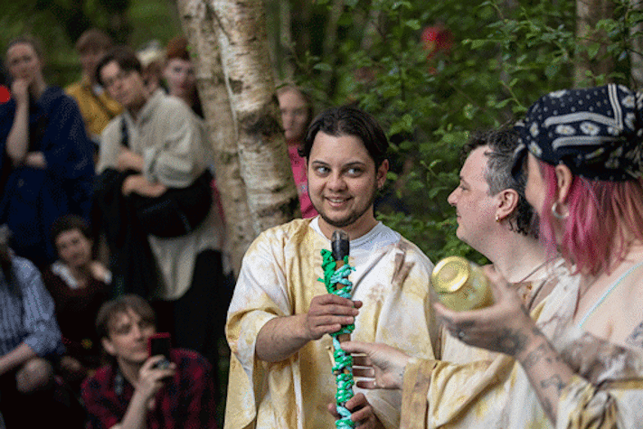 Three people wearing off white cloaks face each other and smile. They pass a stick wrapped in rope to one another.