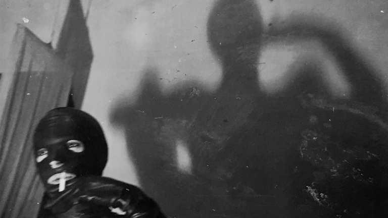 Black-and-white photography. On the bottom left is a person smoking and wearing a latex suit, their shadow casts on to the wall in the background. 