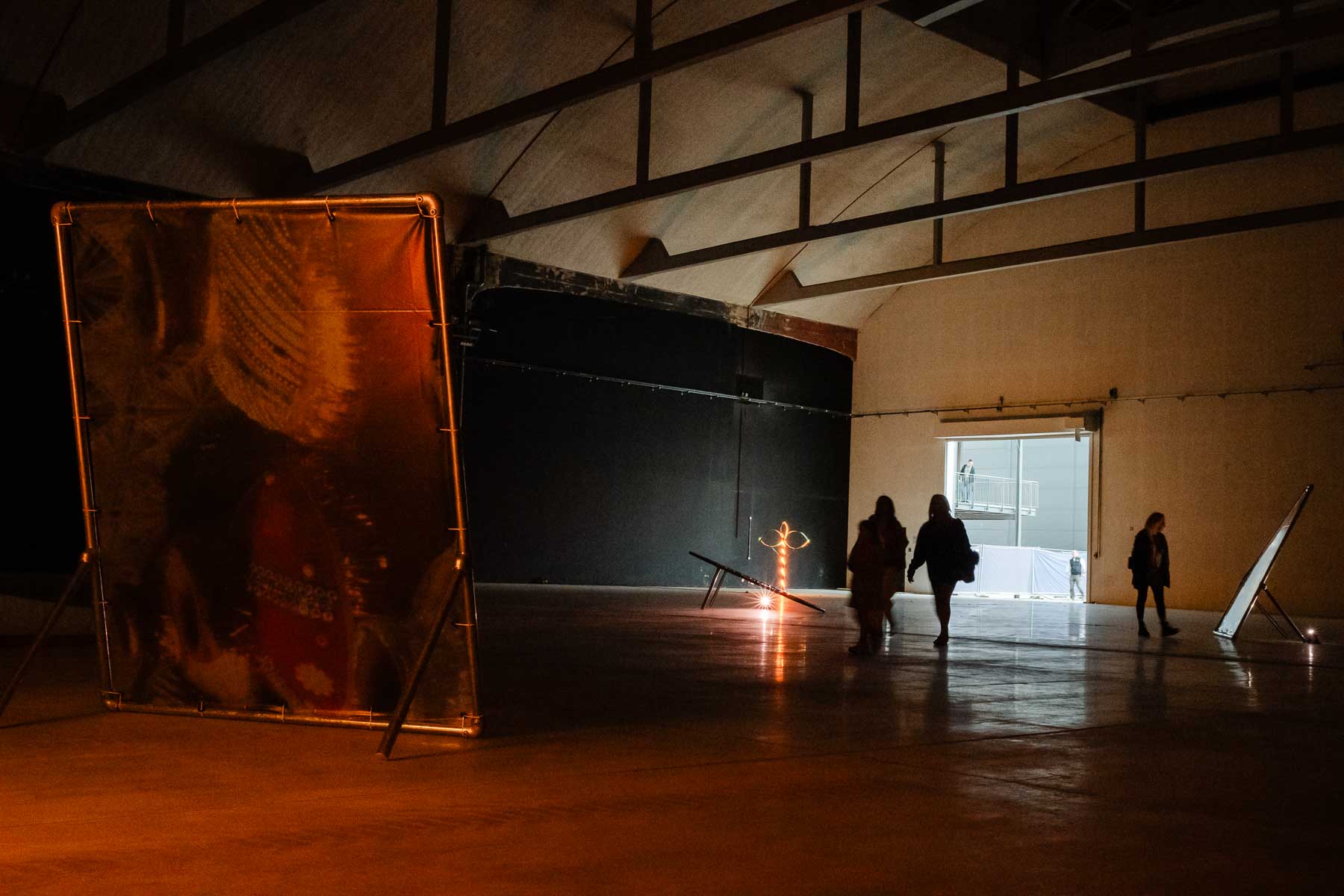 Installation view of a warehouse-style gallery with a large freestanding printed banner in the foreground lit in warm orange tones, a small illuminated sculpture on the floor, and visitors walking through the dimly lit space towards a bright doorway.