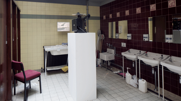 In a bathroom, with bathroom stalls on the left side, with a chair placed in front of the middle stall. There is a tall plinth carrying a projector on it, in the centre of the room. in front of which, is a sink, and able the sink is a small projector screen, displaying an image. On the right side, there is one short sink and two mid height sinks. There are mirrors placed on top of the sinks. 