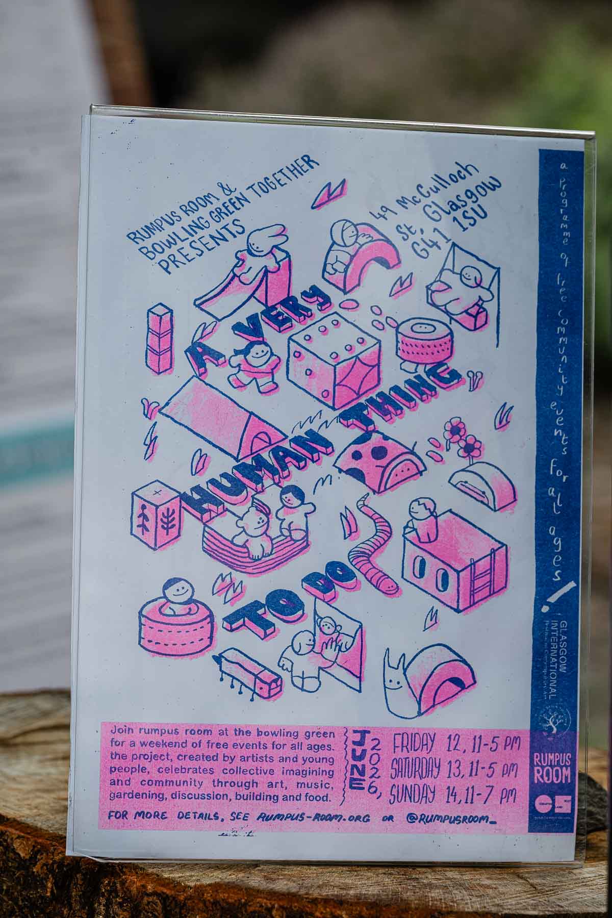 A riso-printed event poster titled "A Very Human Thing To Do" in pink and blue, illustrated with playful icons of play equipment, figures and a snail, propped on a tree stump outdoors.