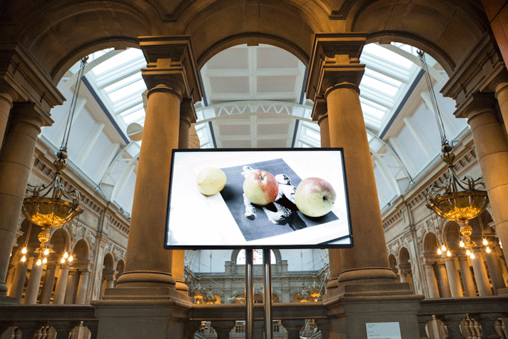 Large arches and pillars are lit up by lights and hanging chandeliers. A long hall with tall arches running along it can be seen past the balcony. In front of the balcony, at the centre, there is a television screen standing on a stand made up of two metal poles. The screen has the image of three apples, ascending in size, beneath which there is a picture of a person holding what seems to be a stuffed toy.