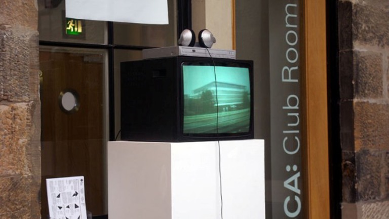 cube monitor atop a plinth. in the background text on the wall reads 'CCA: Club Room'.