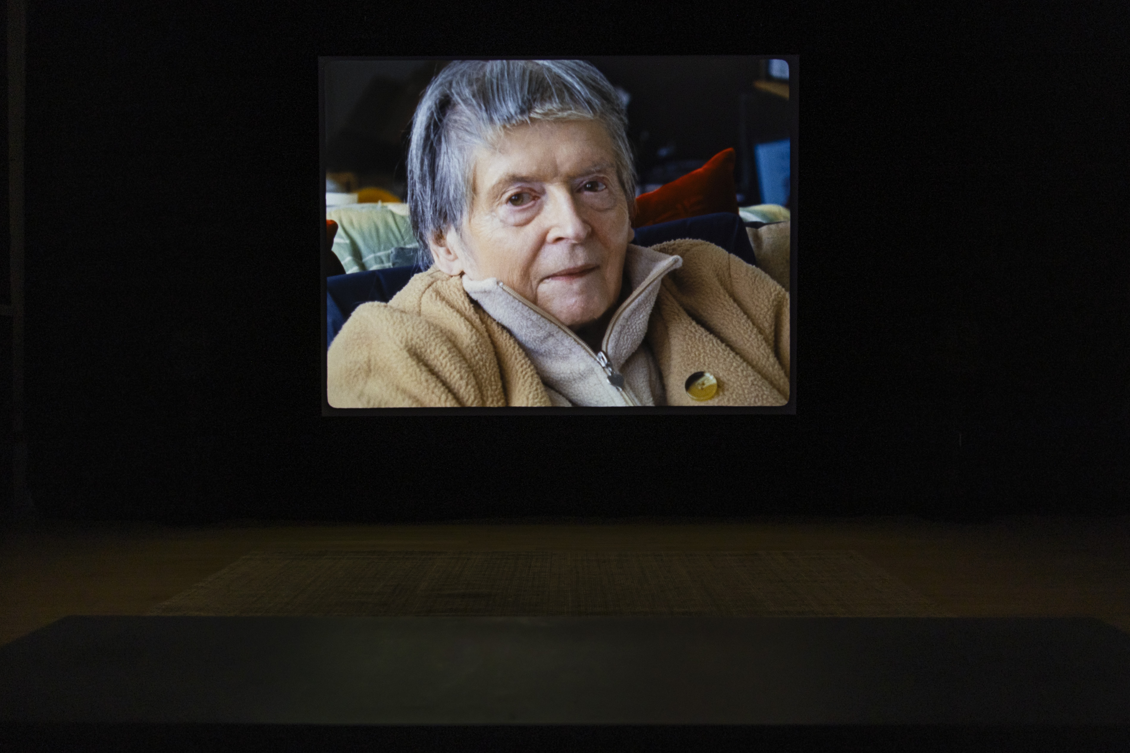 Darkened gallery with a large projected film still showing a close-up portrait of an older woman with grey hair, wearing a beige fleece jacket, seated indoors.