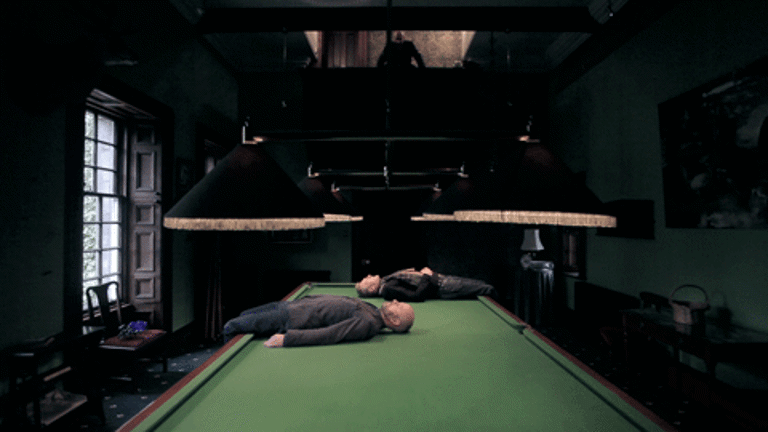 Two men are lying down on the width of a billiards table, with their legs hanging off of the sides. The table is lit up by large lamp lights hanging off the ceiling and a man is looking at the table and the men from the balcony above it.