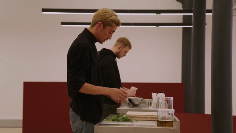 Two white men under fluorescent lights are wearing a black t-shirt and are mixing things like a in a lab experiment. There is a red panel behind them. 