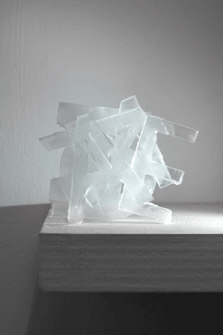 A sculpture made of transparent materials, formed of overlapping letters such as A and E, is placed on a surface attached to the wall.