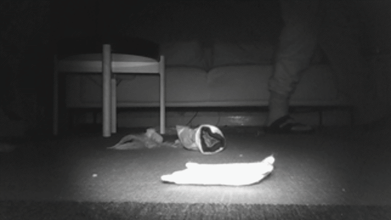 black and white film still image, showing a person sitting on a bed, small bedside table and objects lying on the floor