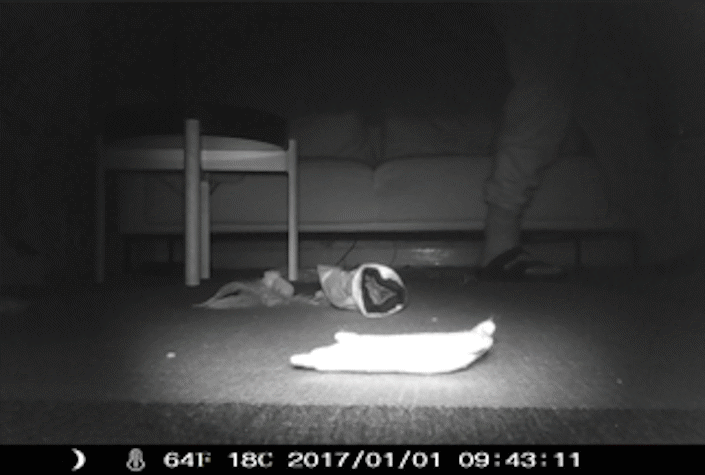 black and white film still image, showing a person sitting on a bed, small bedside table and objects lying on the floor
