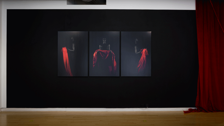 on a black wall at a distance three portrait photographs hang, showing a black figure on a black background holding a bright red drape of fabric