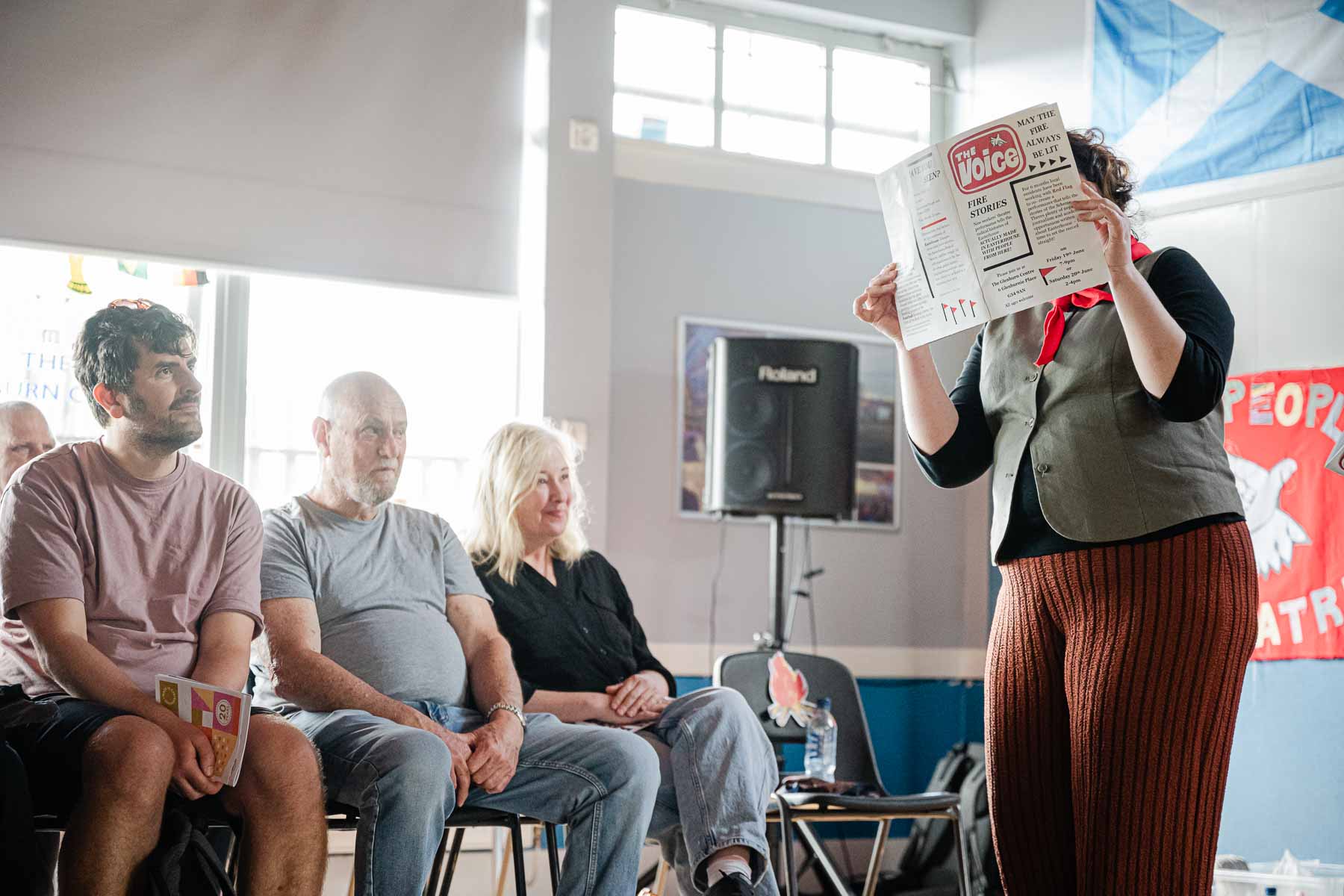 A performer in striped trousers holds up a mock newspaper titled "The Voice" in front of her face, reading from it to a seated audience.