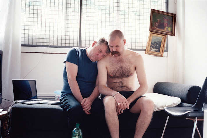 two men sat on a futon. one rests his head not he others shoulder