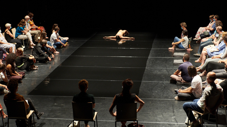  A nude woman with dark hair and olive skin lies face up on a glossy black catwalk stage with her calves tucked underneath her and arms extended outward. An audience gathers around the stage