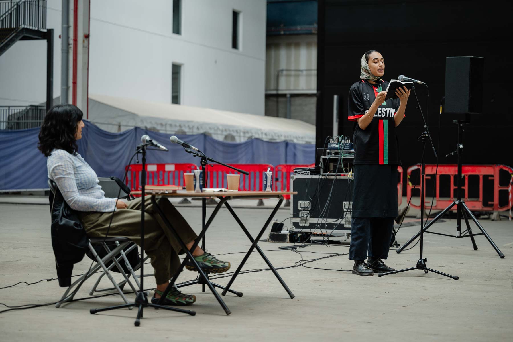 Outdoors, a seated woman listens as another woman stands reading aloud from a book into a microphone, wearing a football shirt reading "Palestine" in red, black and green. A small table with microphones and drinks sits between them, with red barriers and a marquee in the background.