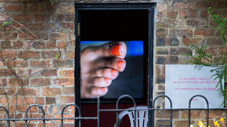 A brick building with flowers in the garden and on the sides of the door. Through the door we can see a video of painted toenails and a white plastic chair. To the right of the open door we can see the title and authors of the exbition in a white sign, pink lettering. 