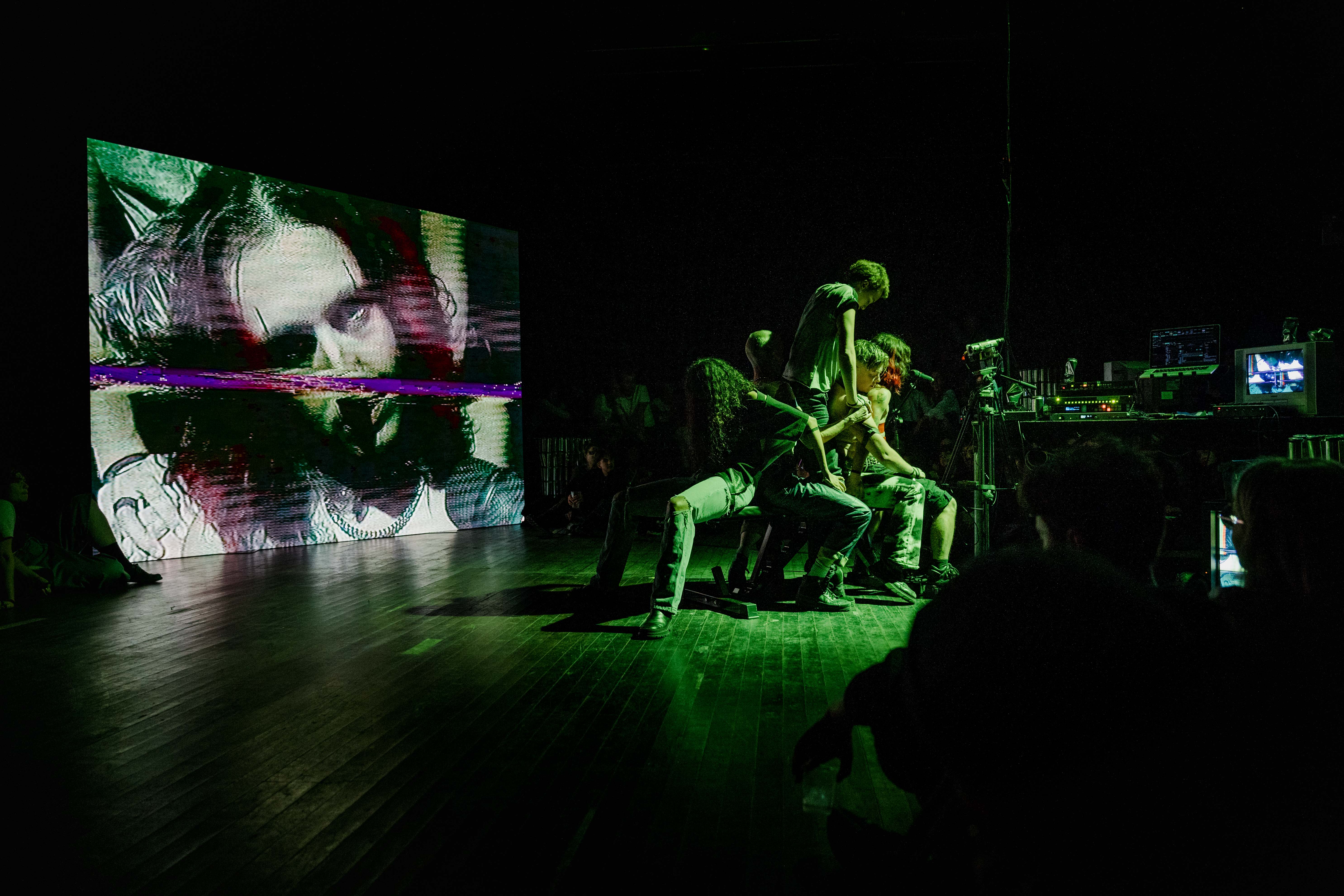 Five dyke-identifying perfomers sit closely together on a weightlifting bench. To the right is a table covered with AV equipment and a camera on a tripod, while to the left a large projection shows another perfomer's head looking back at the group. Audience members sit on the wooden floor around them.
