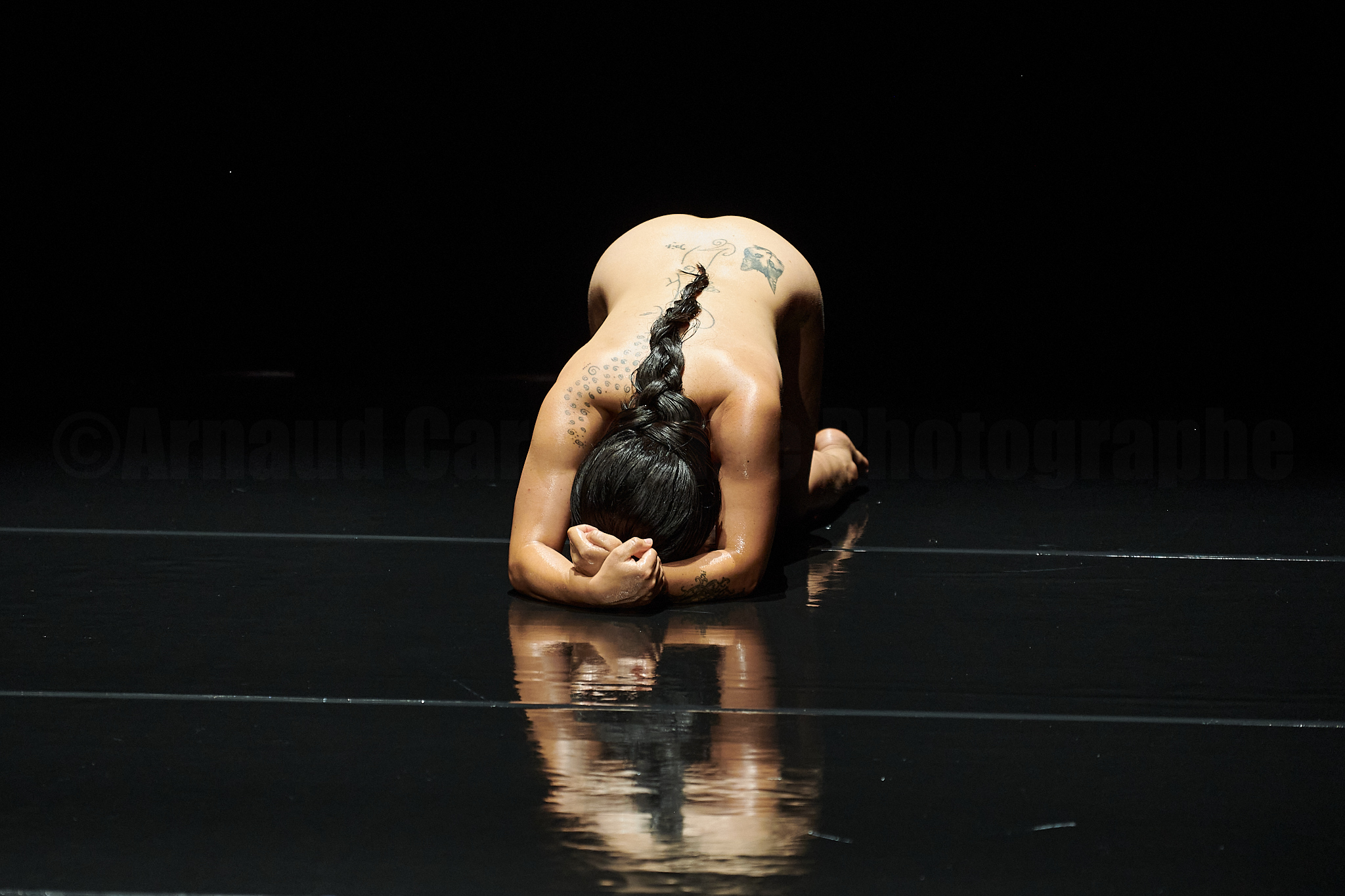 A nude woman with olive skin curls up on her knees on a reflective black surface, her arms wrapped around hear head. A long black braid falls down her back. 