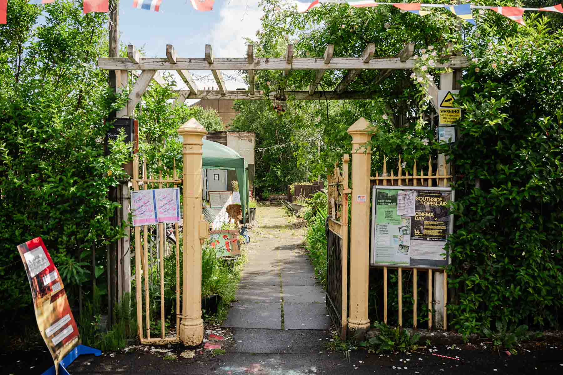 A pergola-covered garden entrance decorated with international flag bunting and climbing roses, leading down a paved path past noticeboards advertising community events.