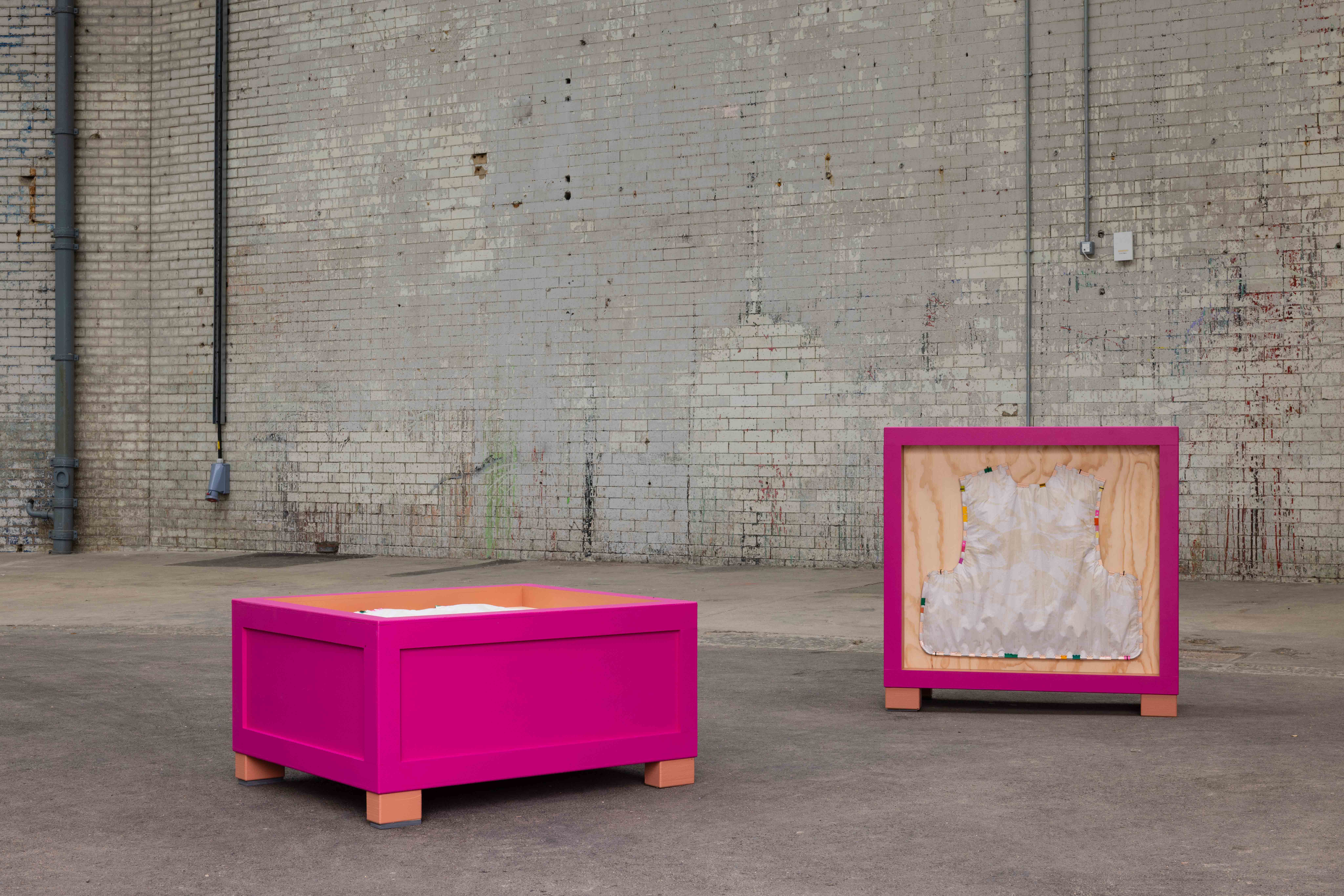 Outdoor installation of two magenta-framed wooden crate sculptures on a paved courtyard against a weathered brick wall, one open box on legs and one displaying a vest-shaped fabric form mounted inside.