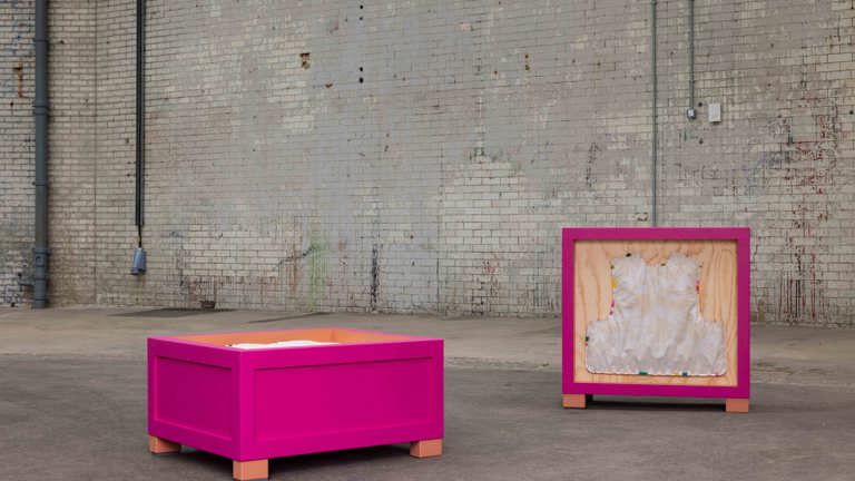 Outdoor installation of two magenta-framed wooden crate sculptures on a paved courtyard against a weathered brick wall, one open box on legs and one displaying a vest-shaped fabric form mounted inside.