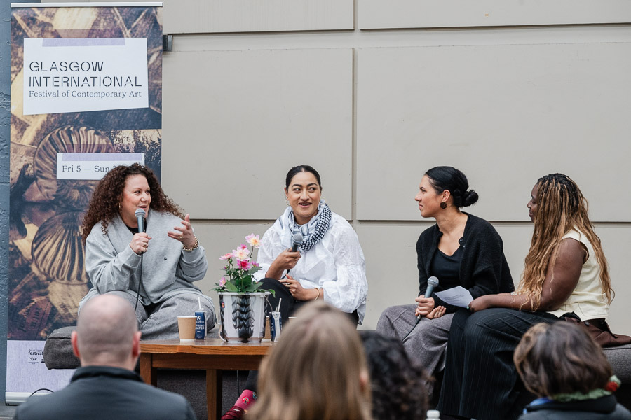 Four women seated on a bench during an outdoor panel discussion, with audience members visible in the foreground. The woman on the left speaks into a microphone mid-gesture, beside a Glasgow International festival banner.