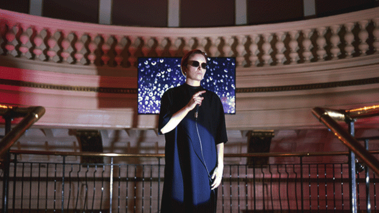 Michelle Hannah is performing in a dark dress and sunglasses, with a microphone in her hand. Behind her, a circular balcony and an image on a screen of purple rain.