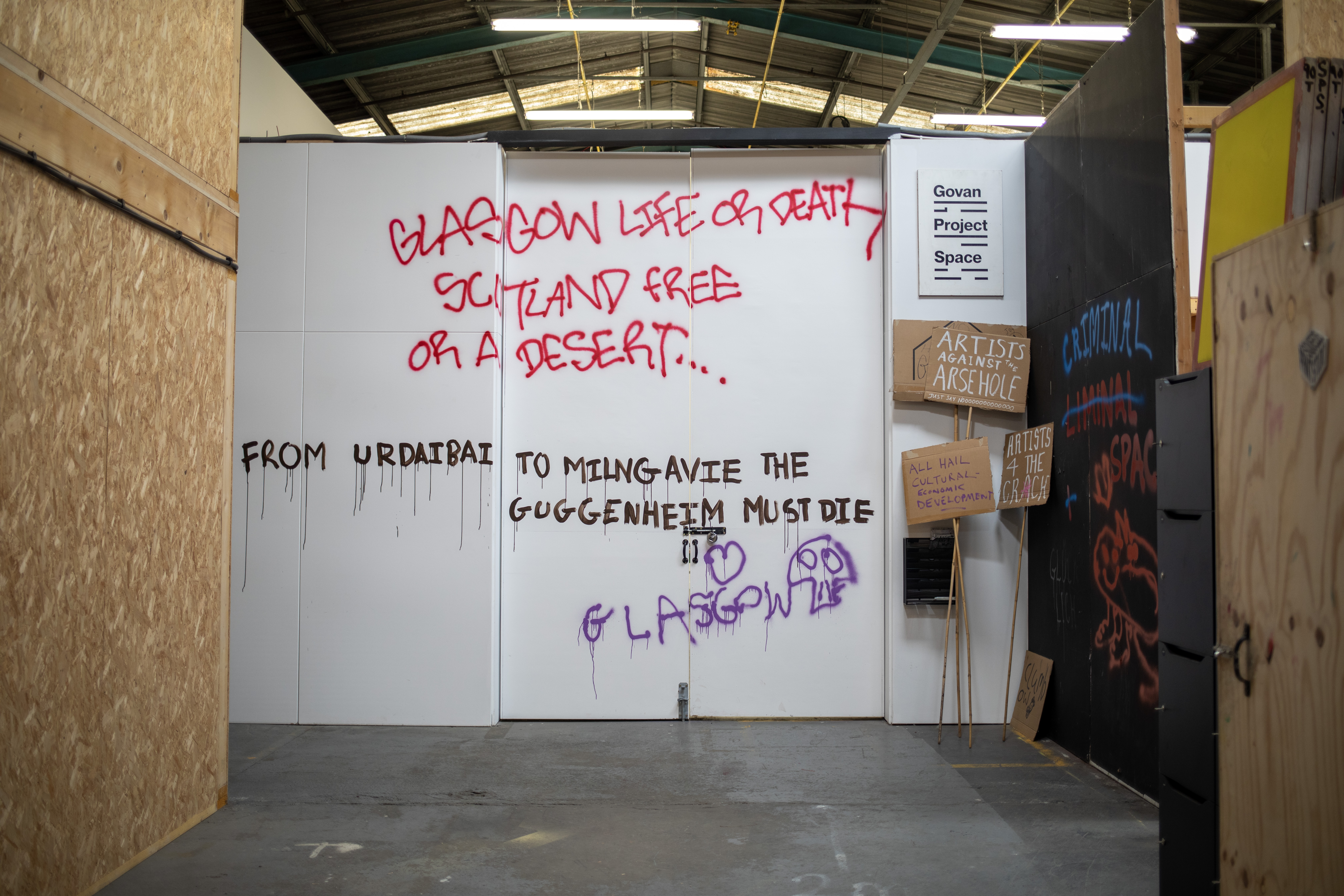 Installation view of the entrance to Govan Project Space, its white doors and walls spray-painted with protest-style graffiti reading "Glasgow Life or Death, Scotland Free or a Desert...", "From Urdaibai to Milngavie the Guggenheim must die" and "Glasgow", with handmade placards reading "Artists Against the Arsehole" and "All Hail Cultural Economic Development" propped against the wall beside a Govan Project Space sign.